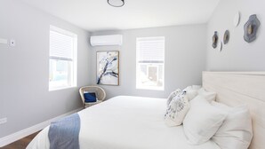 Une chambre avec un grand lit, deux fenêtres équipées de stores, un climatiseur fixé au mur, un fauteuil en osier et un tableau abstrait encadré.
