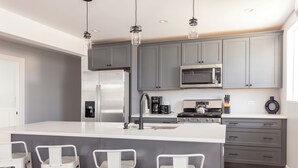 Una cocina moderna con armarios grises, un refrigerador de acero inoxidable, un microondas y una cafetera. Hay tres taburetes blancos en la isla.