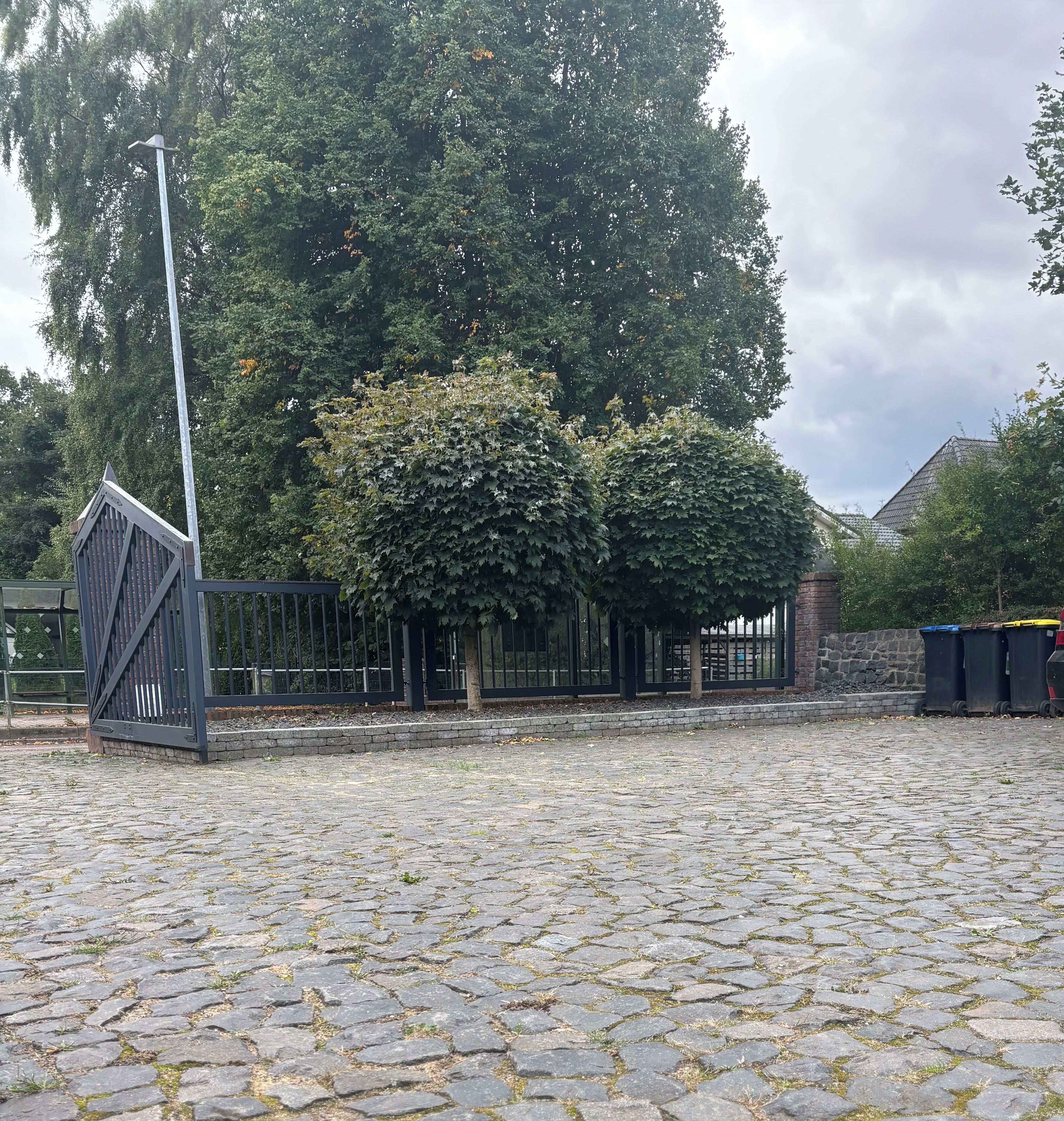 A paved area with a metal fence, trees, and a stone wall.