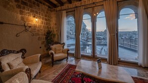 A room with stone walls, a wooden ceiling, a large window with curtains, a sofa, a chair, and a small table. - Majestic Cave House (Nevsehir)