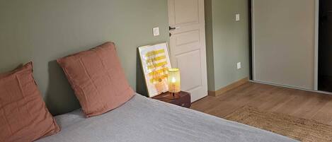 Une chambre à coucher avec un lit, deux oreillers, une table de chevet avec une lampe, une porte et une vue sur un couloir.
