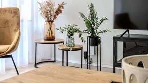 Una sala de estar moderna con una mesa de centro de madera, un sofá, un televisor y tres plantas en macetas sobre mesas auxiliares.