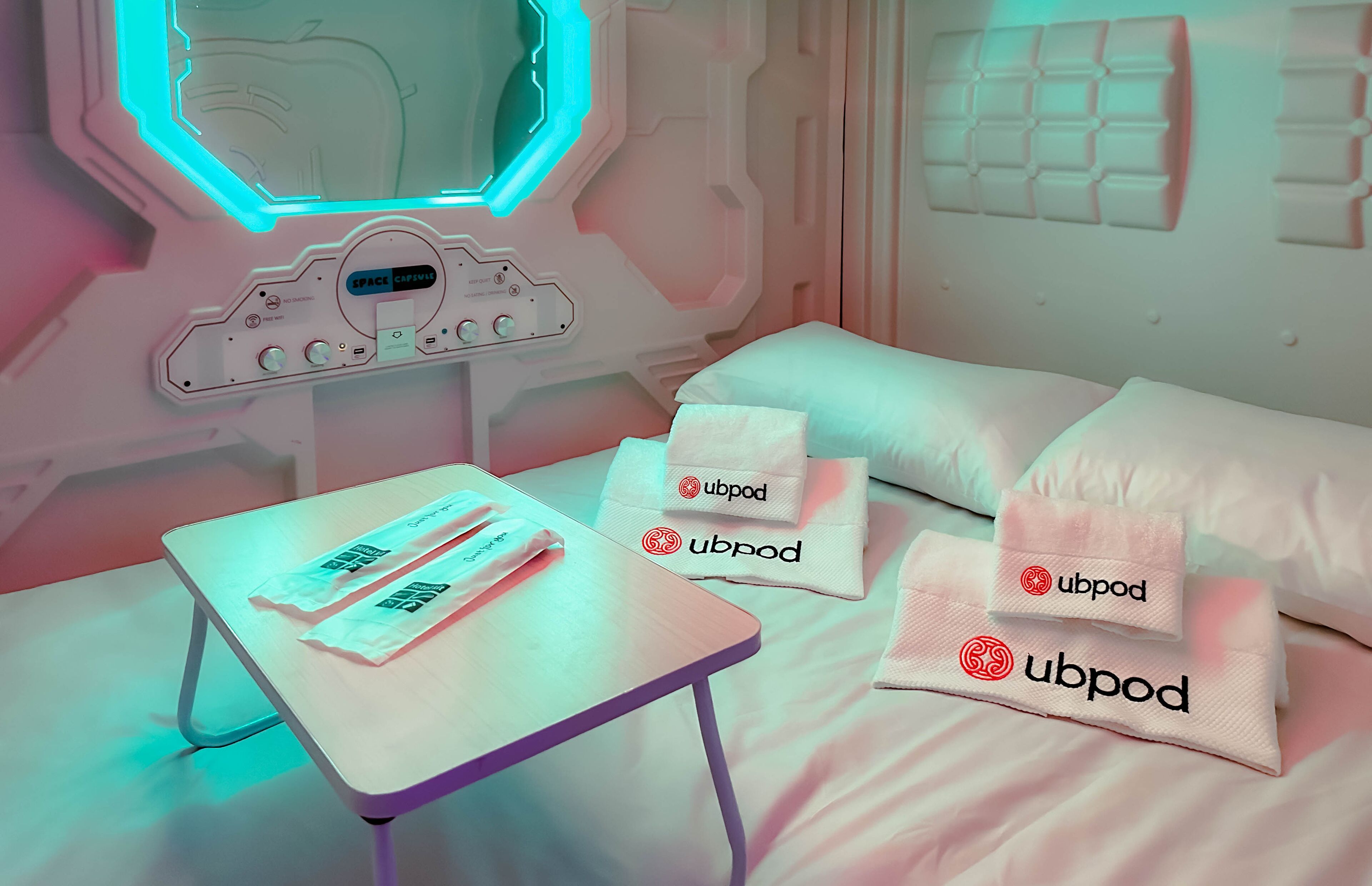 Una habitación con cama, almohadas, una mesa con objetos y un panel de control en la pared.