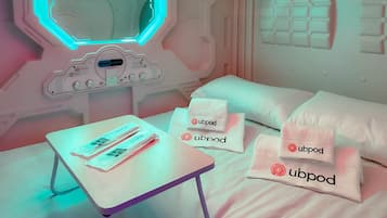 Una habitación con cama, almohadas, una mesa con objetos y un panel de control en la pared.