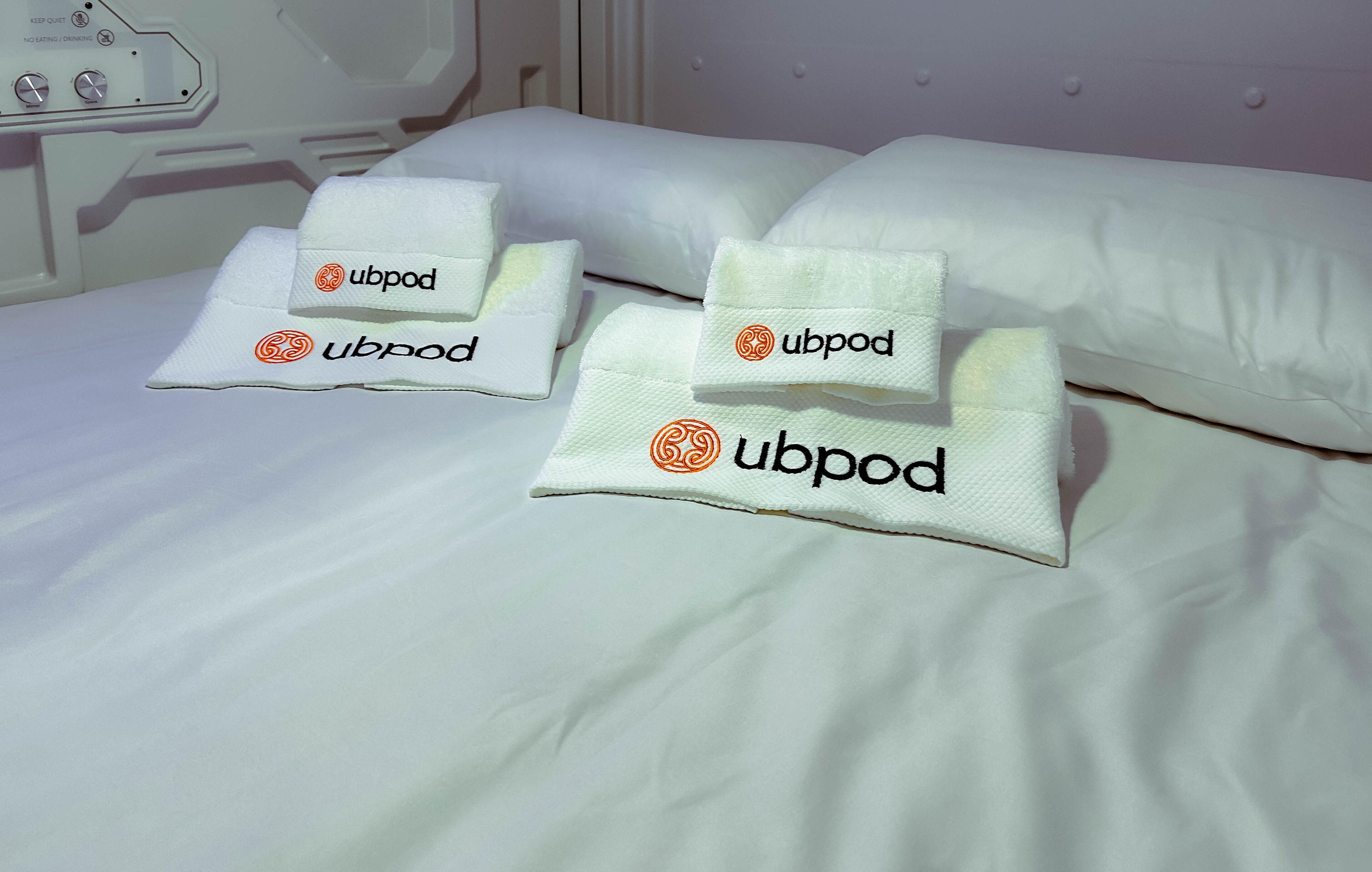 A bed with white bedding and two folded towels with 'ubpod' branding.