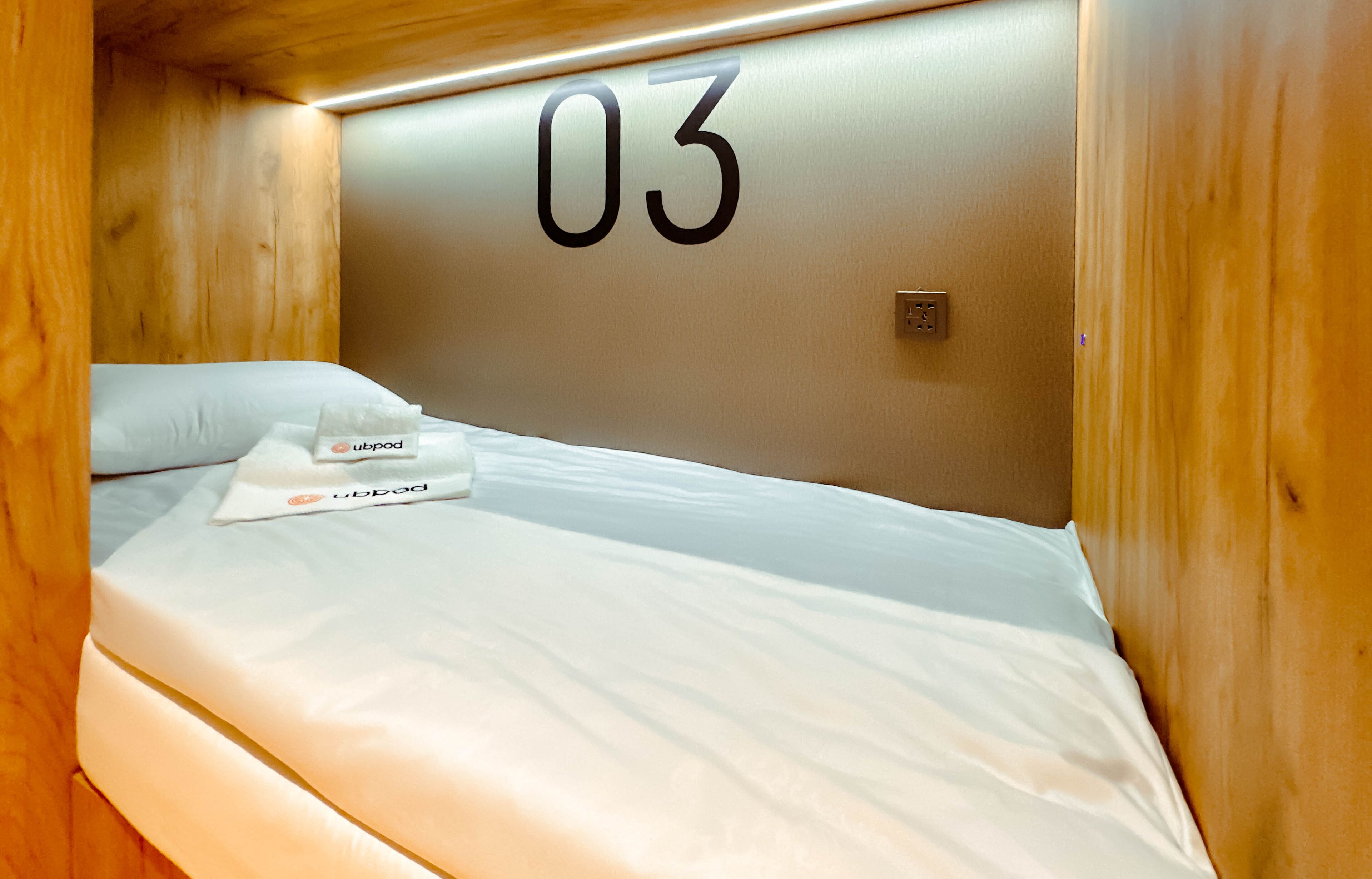 Una cama individual con el número '03' en la pared, una almohada 'ubpod' y una etiqueta que indica 'pod hotel'.