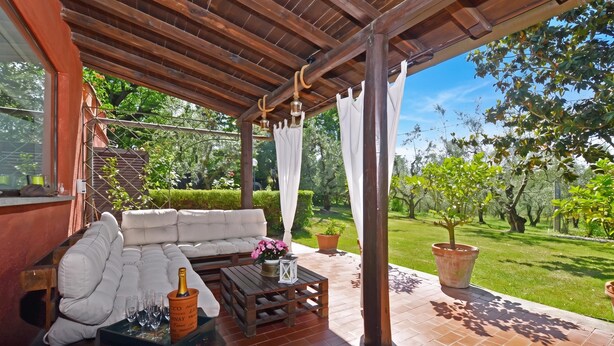 A covered patio with a wooden ceiling, a white cushioned sofa, a wooden coffee table, and a potted plant. - Casale Pontarello (Vetralla)