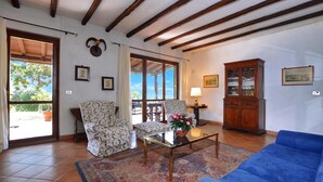 A living room with a blue sofa, patterned armchairs, a glass coffee table, a wooden cabinet, and a view through a glass door. - Casale Pontarello (Vetralla)