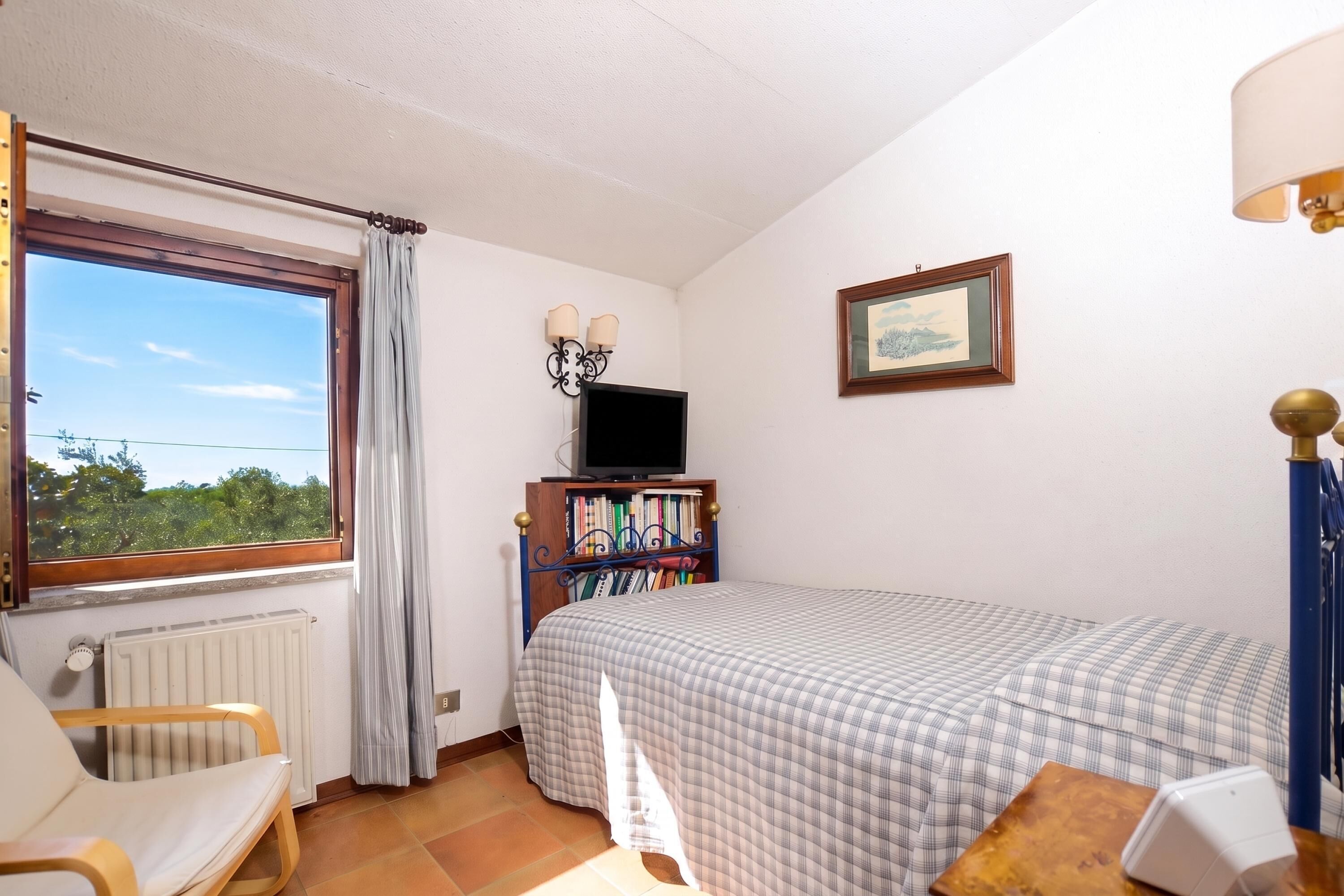 Une chambre à coucher avec un lit, une chaise, un radiateur, une fenêtre avec des rideaux, une bibliothèque, un téléviseur et un tableau encadré au mur.