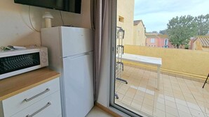 Et værelse med et fladskærms-TV monteret på væggen, et hvidt køleskab, en mikrobølgeovn og et lille bord med en stol uden for en skydedør.