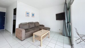 A modern living room with a sectional sofa, a wooden coffee table, a flat-screen TV mounted on the wall, and a large window with curtains. - Air-conditioned apartment for 4 with pool, terrace and sea view (Le Barcarès)