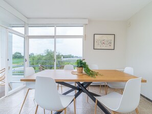 A dining area with a large table, white chairs, a potted plant, and a framed artwork on the wall.