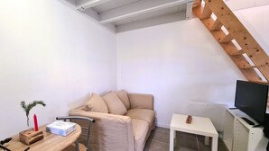 Een kleine, goed verlichte kamer met een beige bank, een kleintje tafeltje, een televisie op een meubel en een houten ladder tegen de muur.