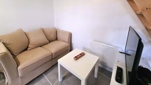 Een kleine, moderne woonruimte met een beige bank, een witte salontafel, een flatscreen televisie en een afstandsbediening op de vloer.
