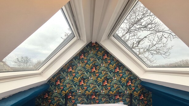 Een kamer met een schuine kap, een bed geplaatst onder twee dakramen, en een kamerwand met een patroon.
