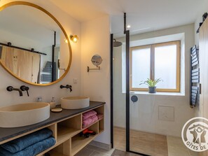 Une salle de bain moderne avec un grand miroir rond, deux vasques, une douche et une fenêtre agrémentée d’une plante.
