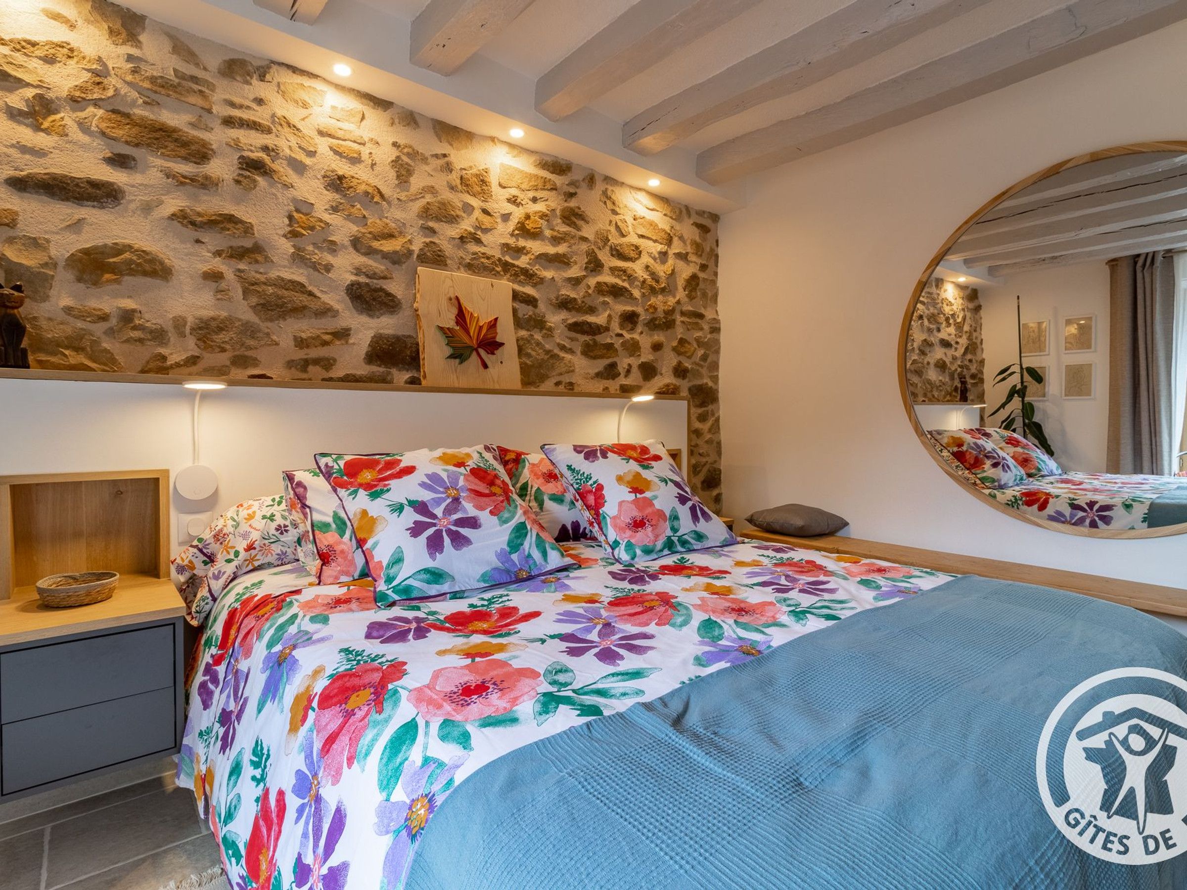 A bedroom with a stone wall, a bed with floral bedding, a bedside table with a bowl, and a round mirror on the wall.