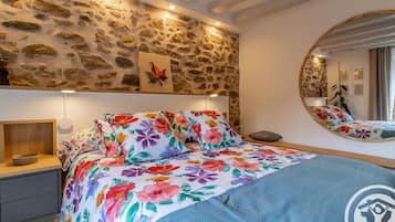 A bedroom with a stone wall, a bed with floral bedding, a bedside table with a bowl, and a round mirror on the wall.