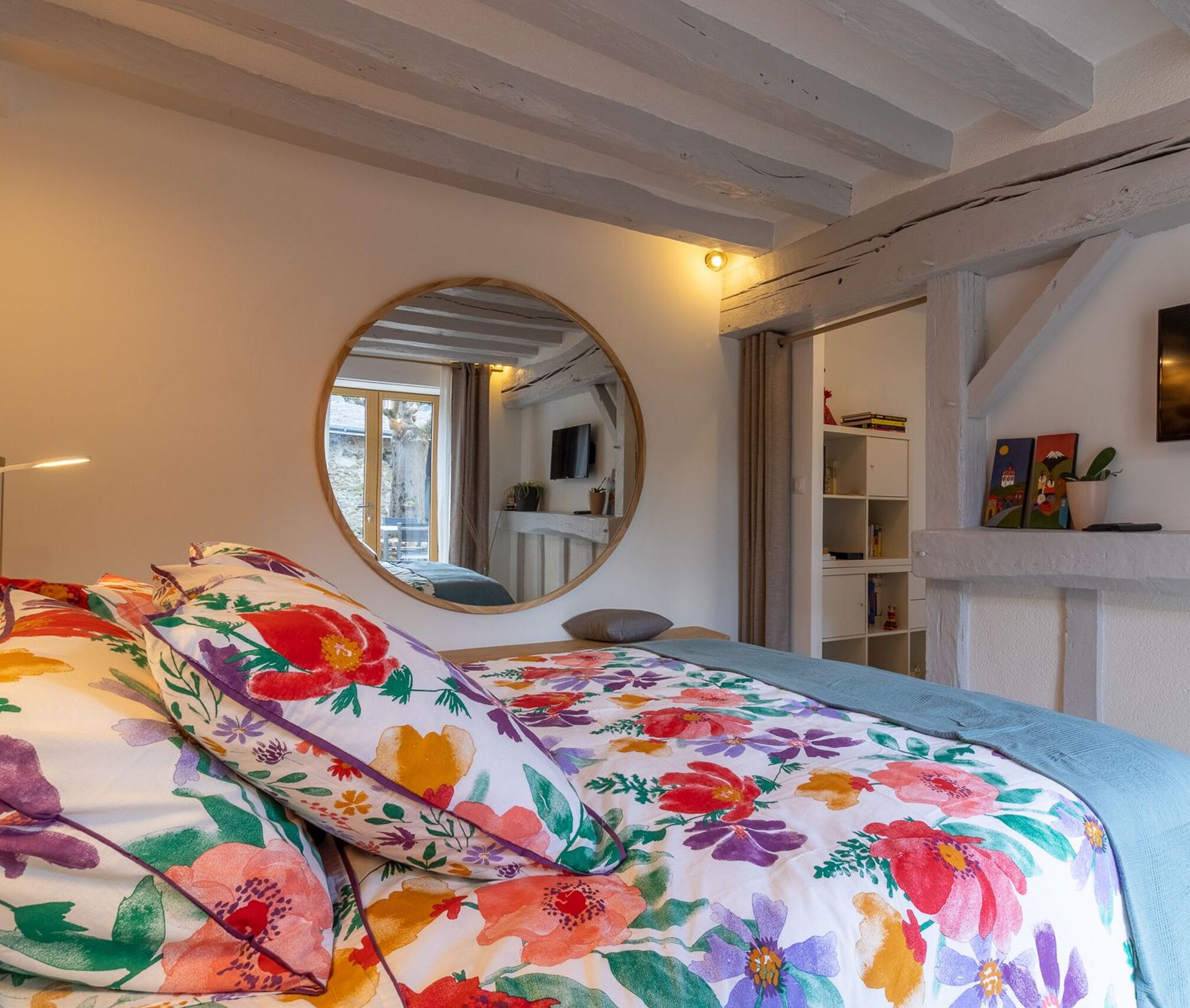 A bedroom with a bed, a round mirror, a bookshelf, and a window.