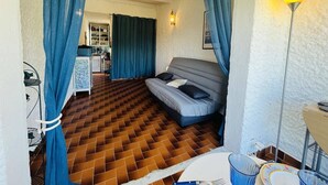 A compact room with a patterned floor, a bed with pillows, a desk with a lamp, and a kitchenette in the background.