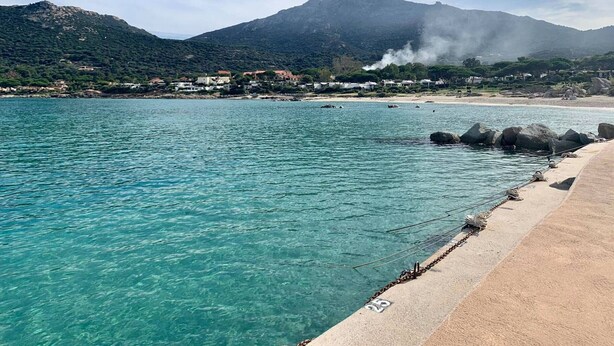 A coastal scene with clear turquoise waters, a sandy beach, and a backdrop of mountains.