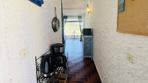 A narrow hallway with white walls, a tiled floor, a coffee machine, a hanging pot, and a bulletin board with posters.