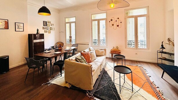 Un salon chaleureux avec un canapé, une table à manger et des chaises. On y trouve des fenêtres avec des volets, un luminaire suspendu et un tapis sur le sol.