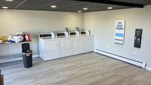 Une buanderie avec une rangée de machines à laver et de sèche-linge, une poubelle et un distributeur de savon fixé au mur.