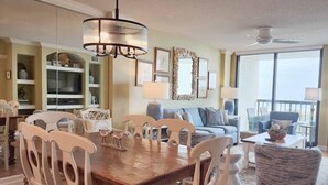 A spacious living room with a wooden dining table, white chairs, a blue sofa, and a balcony with a view. - Crescent D4E Litchfield Beach (Pawleys Island)