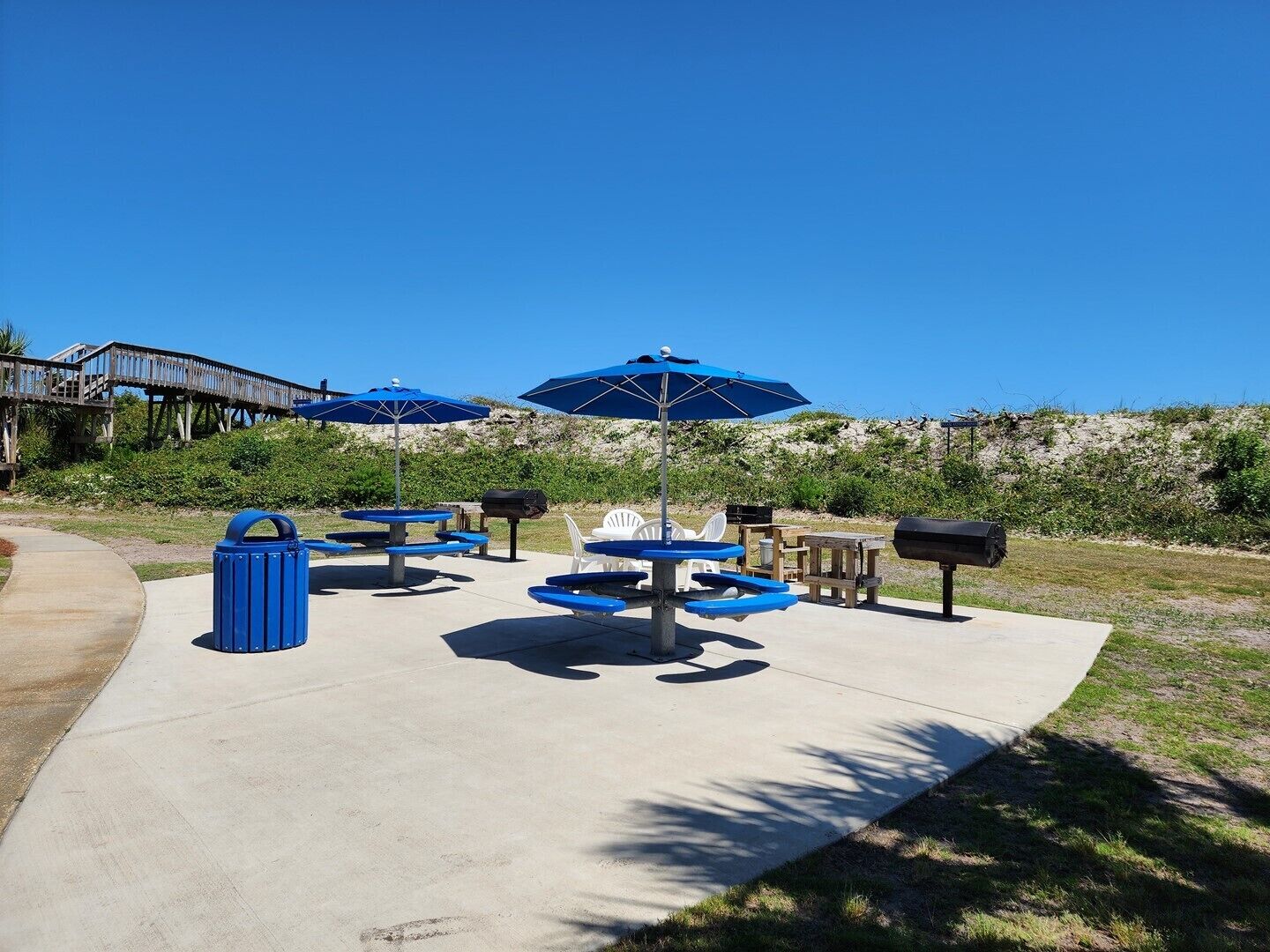 Un espace extérieur aménagé avec des tables et des parasols bleus, une terrasse en bois et une vue sur une plage et une promenade.
