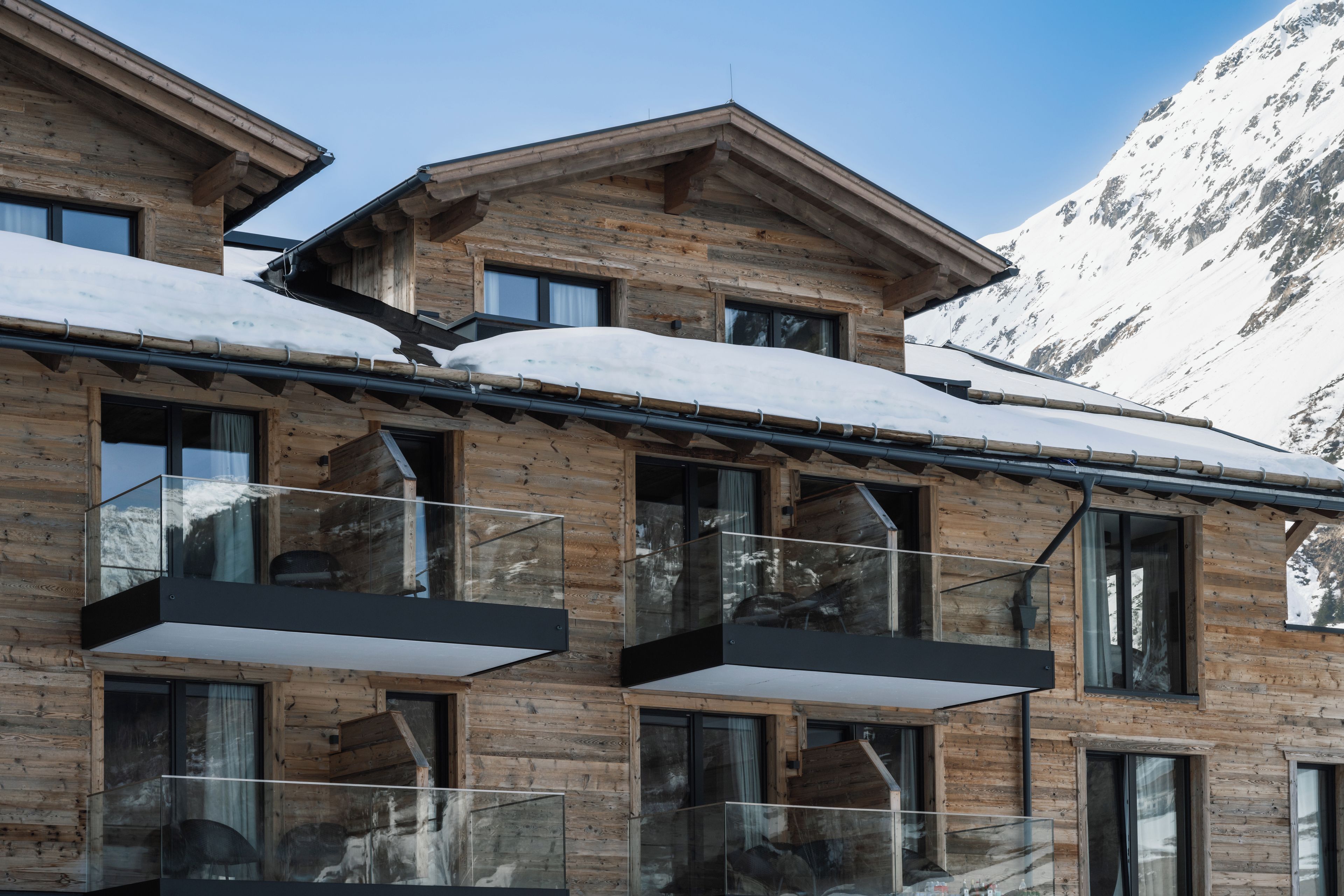 A wooden chalet with multiple balconies, large windows, and a snowy mountain backdrop.