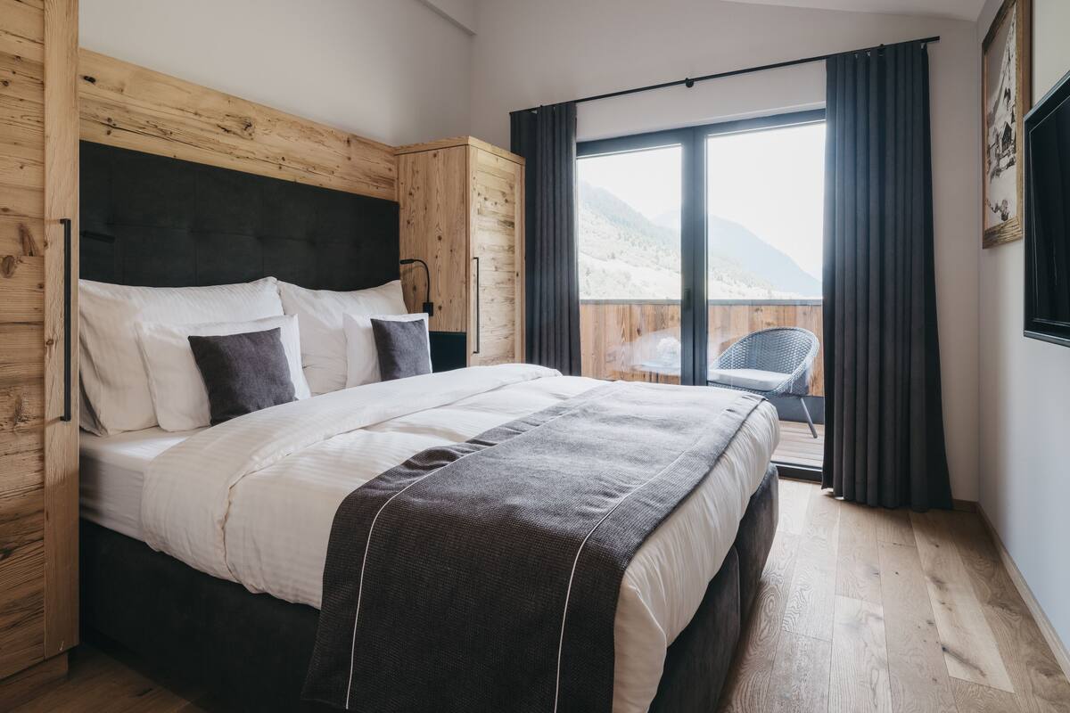 Une chambre avec une tête de lit en bois, un grand lit avec une literie blanche et foncée, une vue sur un balcon avec une chaise, et un paysage montagneux en arrière-plan.