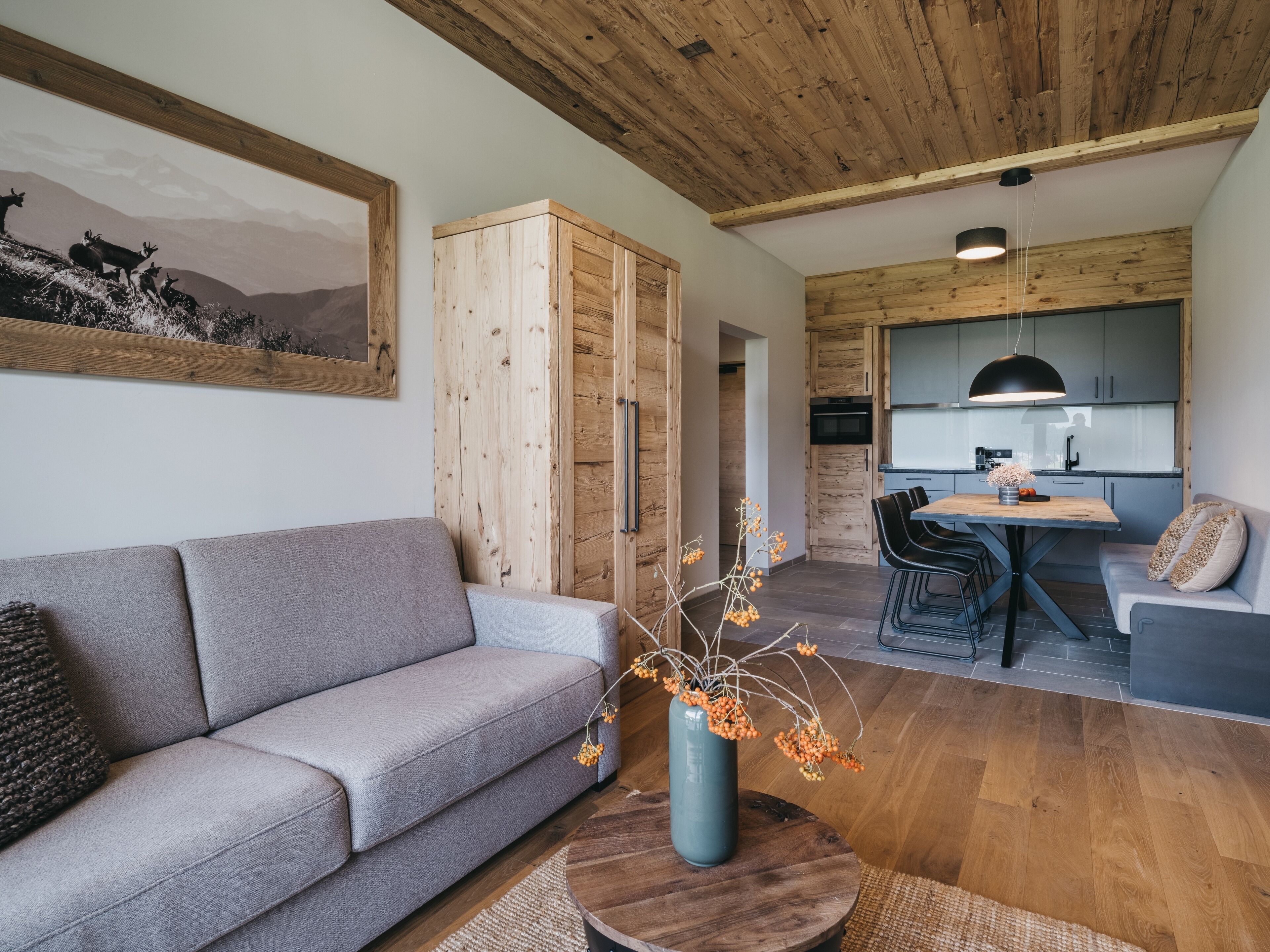 Ein modernes Wohnzimmer mit grauem Sofa, einem Holzesstisch und einer Küchenzeile mit grauen Schränken und einer Pendelleuchte.