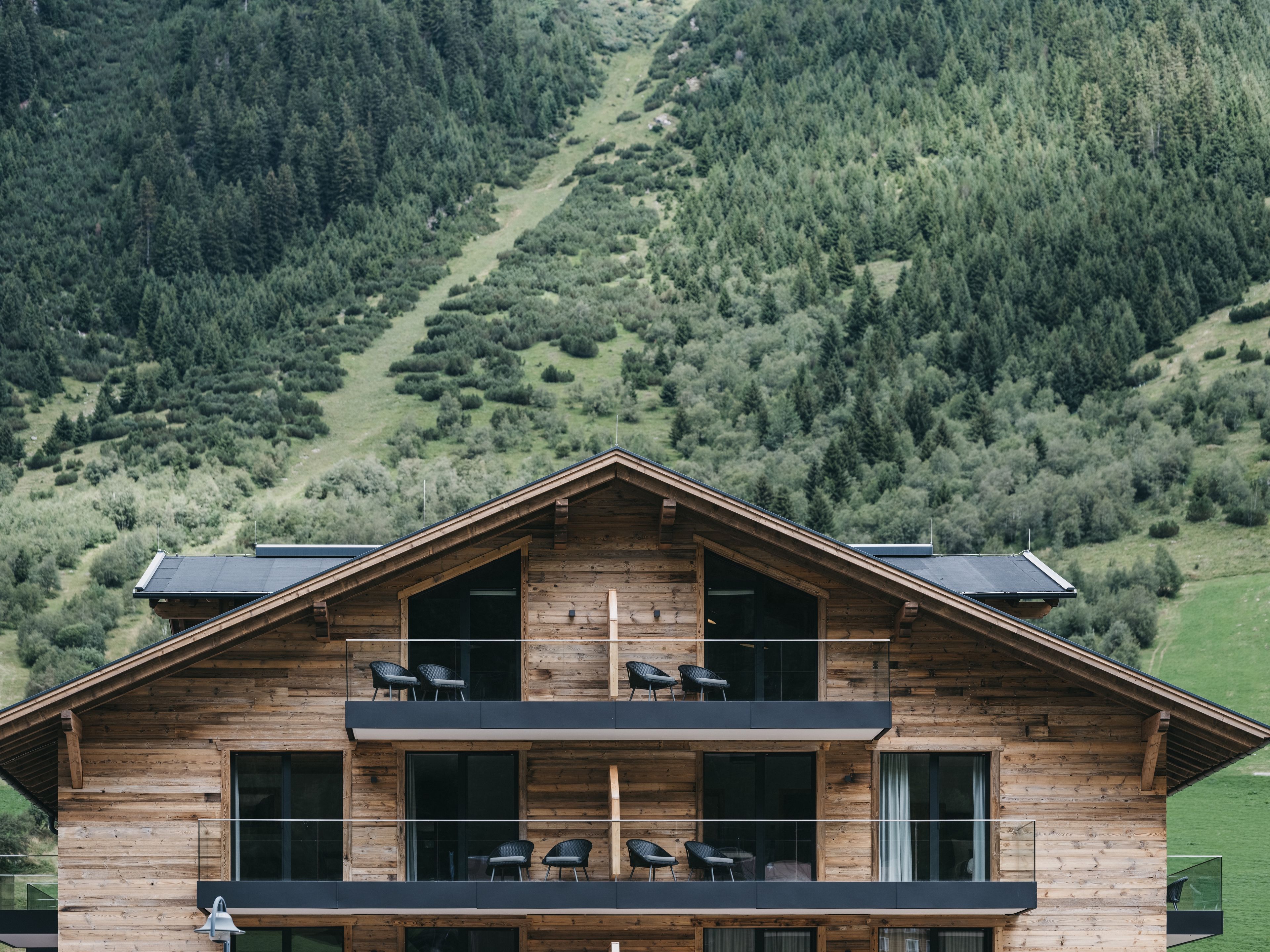 A two-story wooden cabin with a balcony, large windows, and a view of a forested hillside.