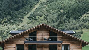 A two-story wooden cabin with a balcony, large windows, and a view of a forested hillside.