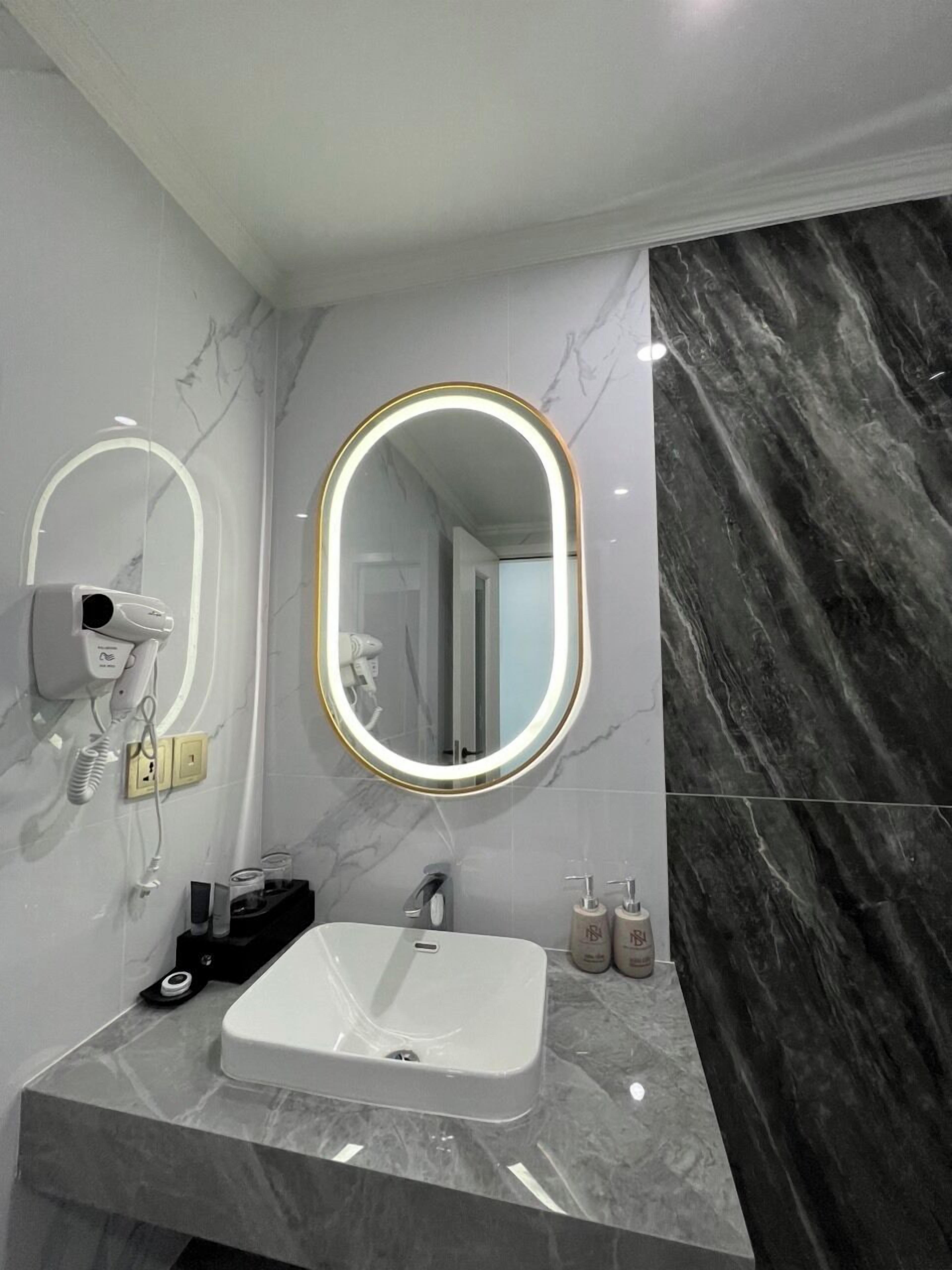 Un baño moderno con un lavamanos blanco, un espejo ovalado grande con marco dorado y un secador de pelo montado en la pared.