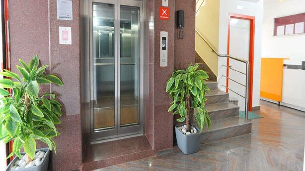 Een lift met glazen deuren, een rood rookverbod-bord, een beveiligingscamera en een telefoon aan de muur.