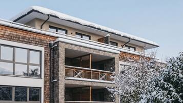 A multi-story building with a balcony on the ground floor, surrounded by snow-covered vegetation.