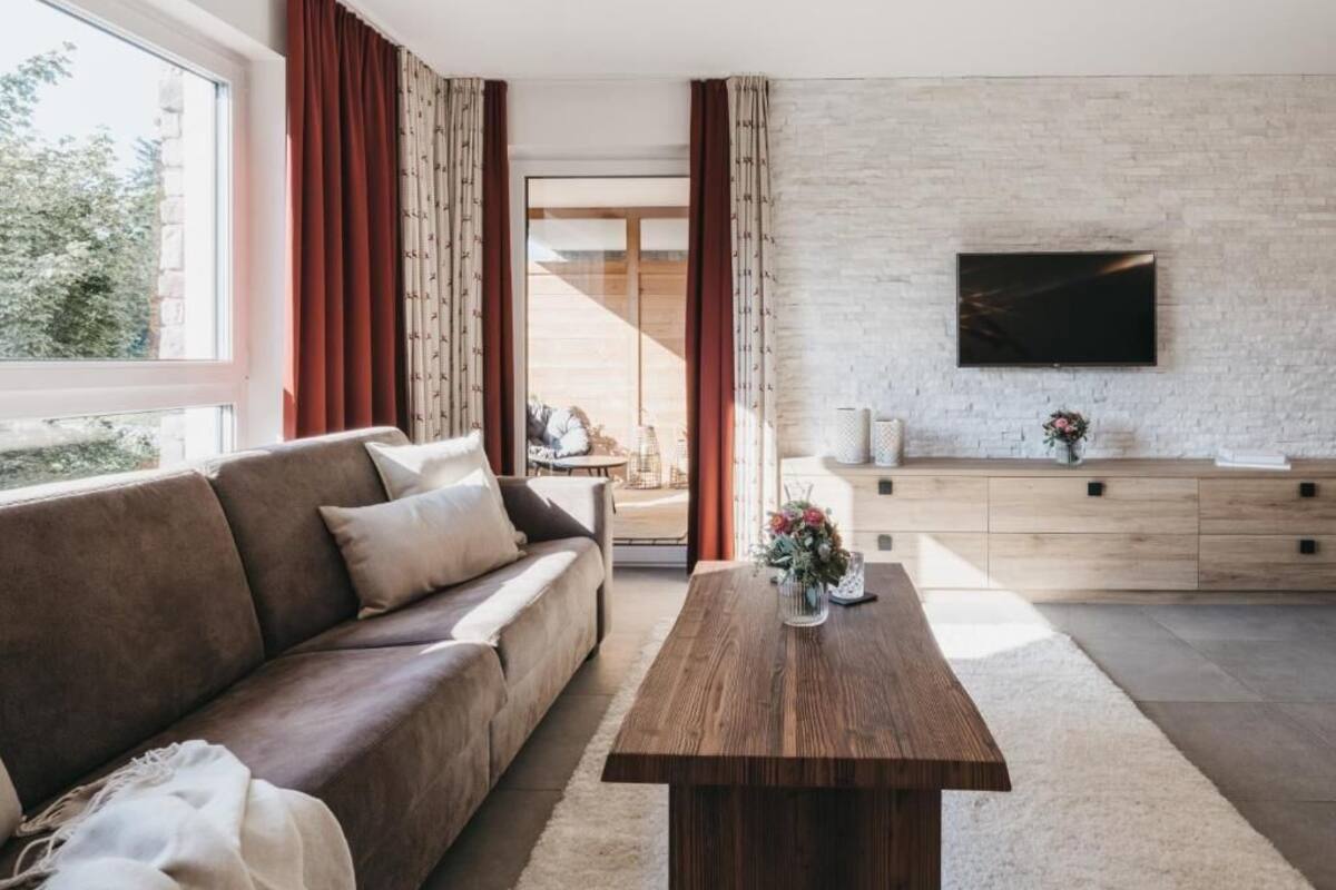 Una sala de estar moderna con un sofá, una mesa de centro de madera, un televisor montado en una pared de ladrillo y vistas al exterior.