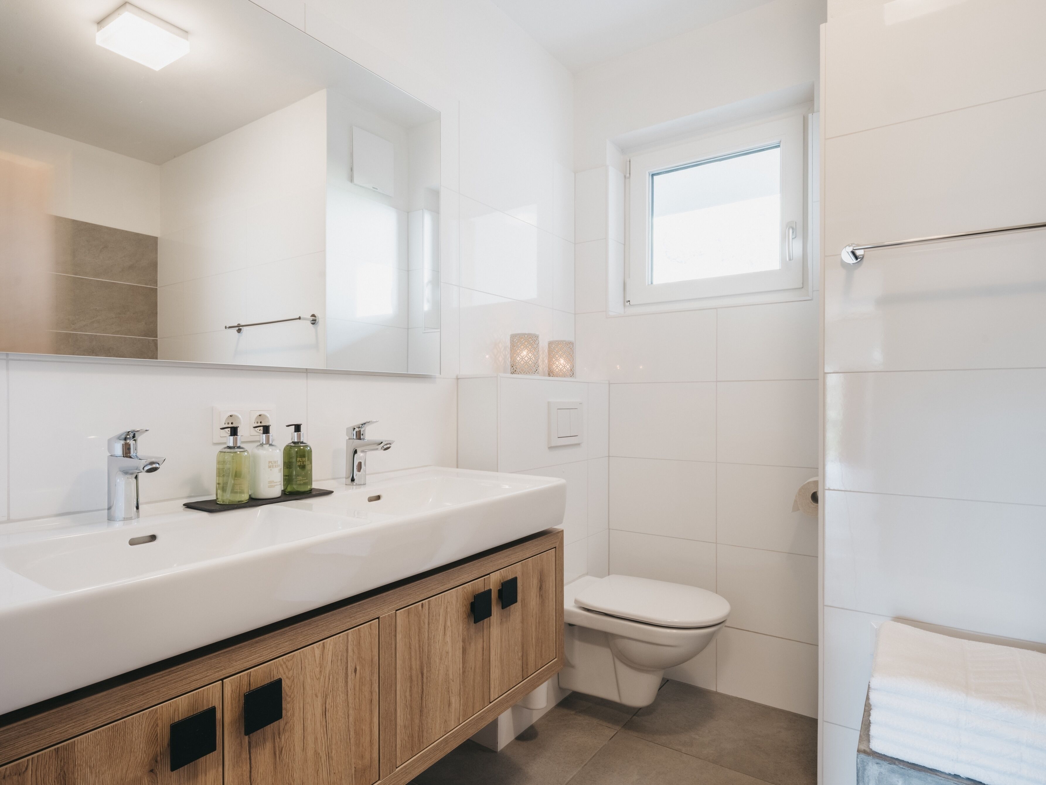 A modern bathroom with a white vanity, two sinks, a toilet, and a window.