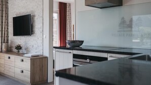 A modern kitchen with a flat-screen TV mounted on a brick wall, a large island with a bowl, and a view of Village Park Suites. - Village Park Suites (Zell am See)
