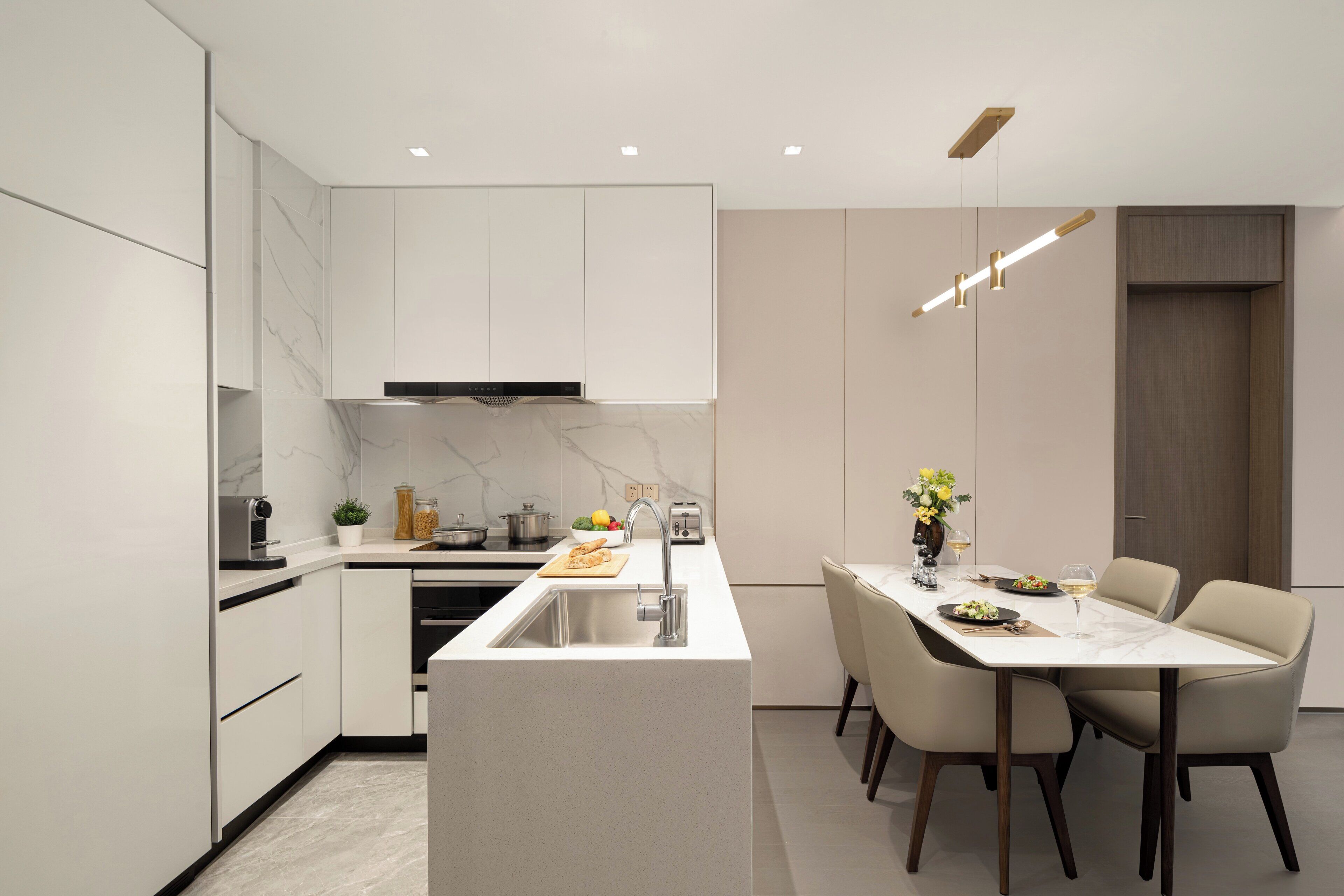 Cozinha moderna com armários brancos, painel de mármore e uma ilha central com pia. Há uma área de refeições com mesa e cadeiras, e um vaso com flores na ilha.
