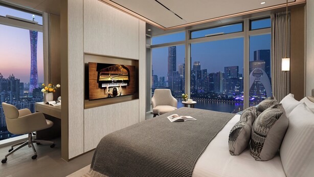 A modern hotel room with a large bed, a desk, and a TV. The room has a city view through large windows.