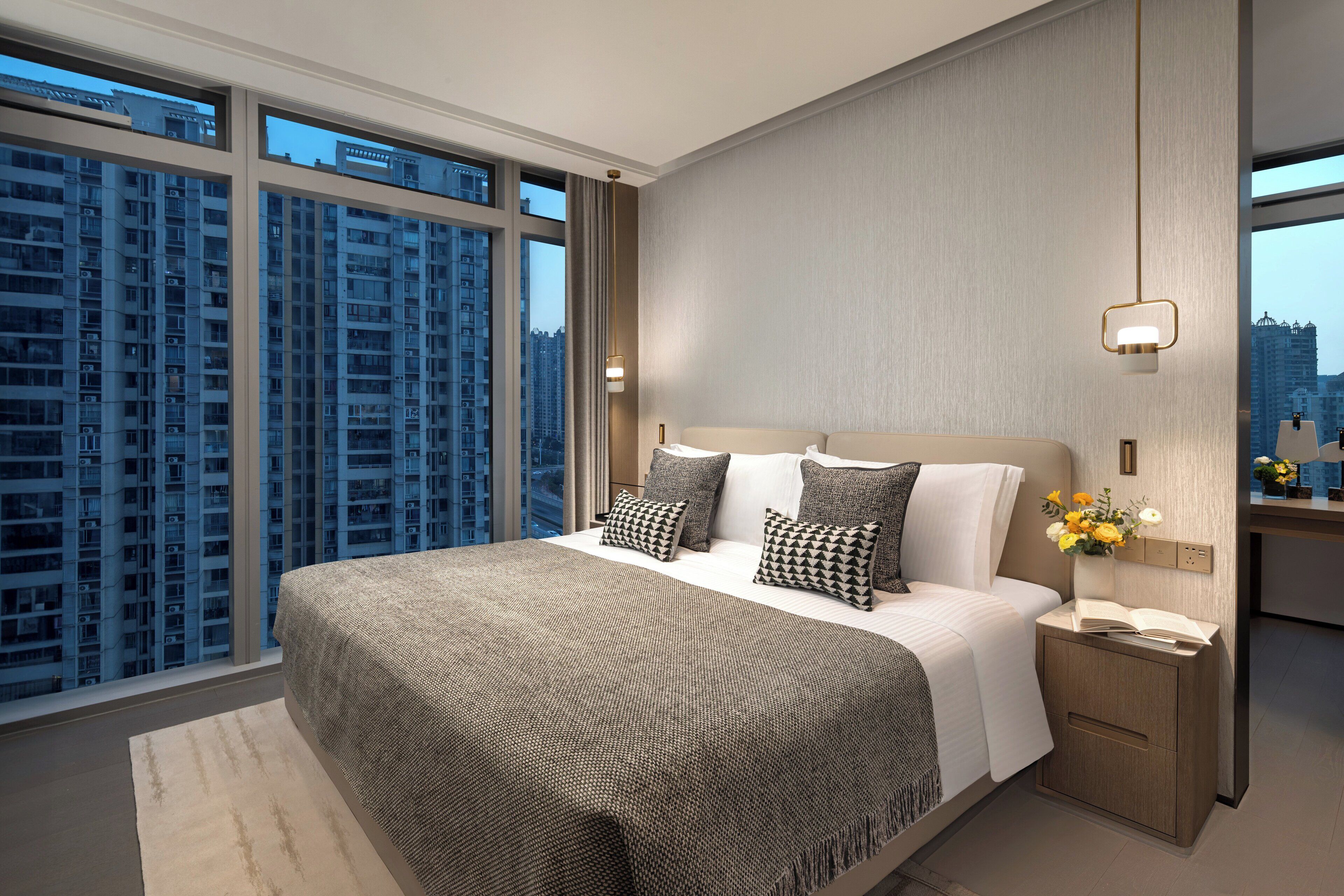 Quarto de hotel moderno com uma cama grande, uma mesa de cabeceira, uma luminária e vista para edifícios altos.