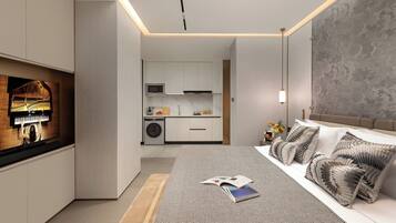 A modern bedroom with a large bed, patterned pillows, a bedside table with a book, and a view into the adjacent kitchen area.