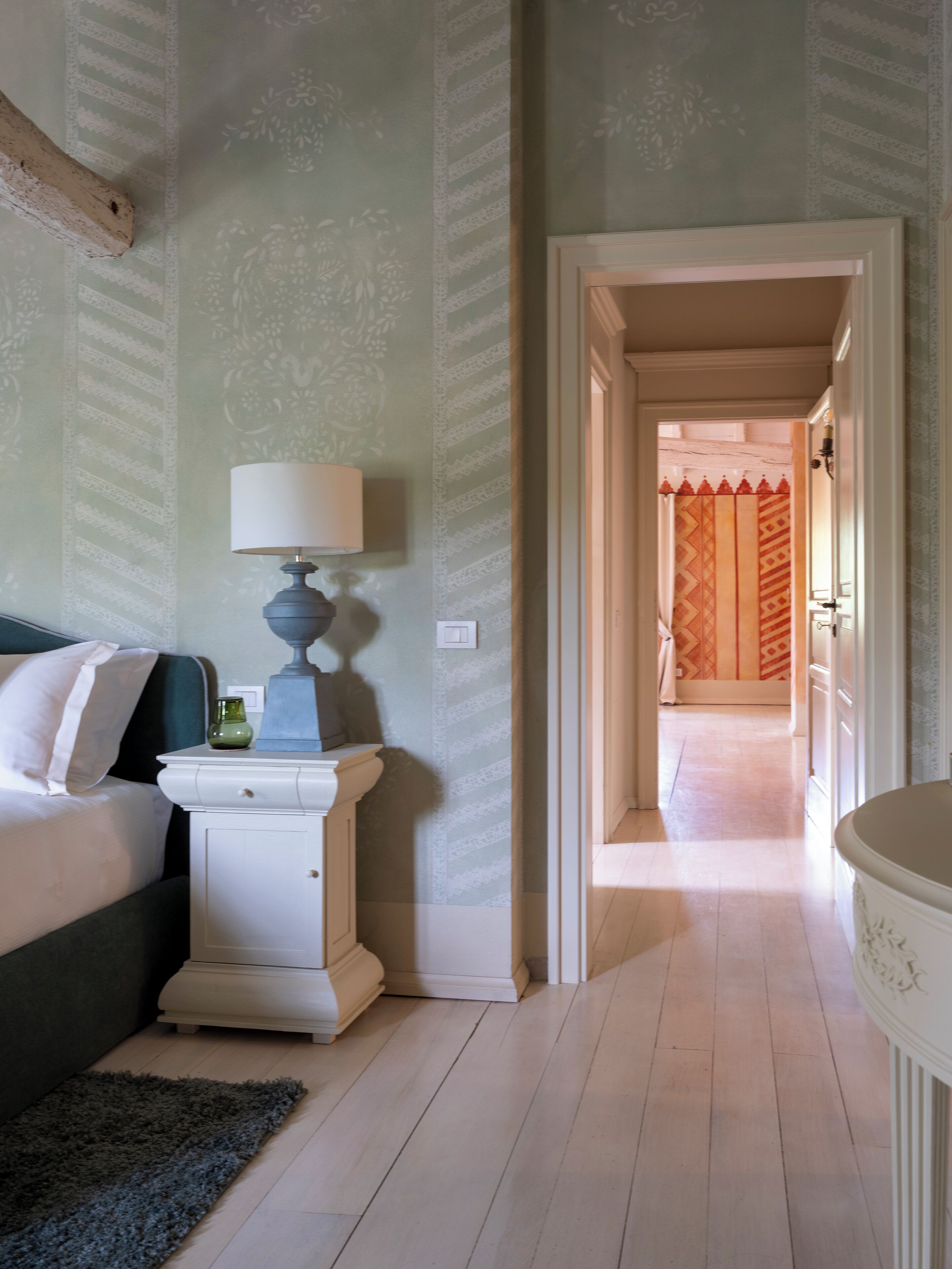 Une chambre à coucher avec un lit, une table de chevet, une lampe et une vue sur une pièce adjacente, séparée par un rideau à motifs.