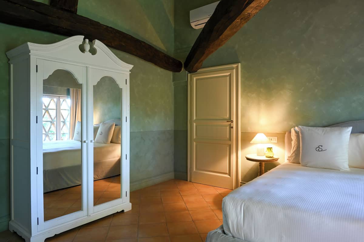 A bedroom with a large white wardrobe, a bed with white bedding, a nightstand with a lamp, and a door leading to another room.