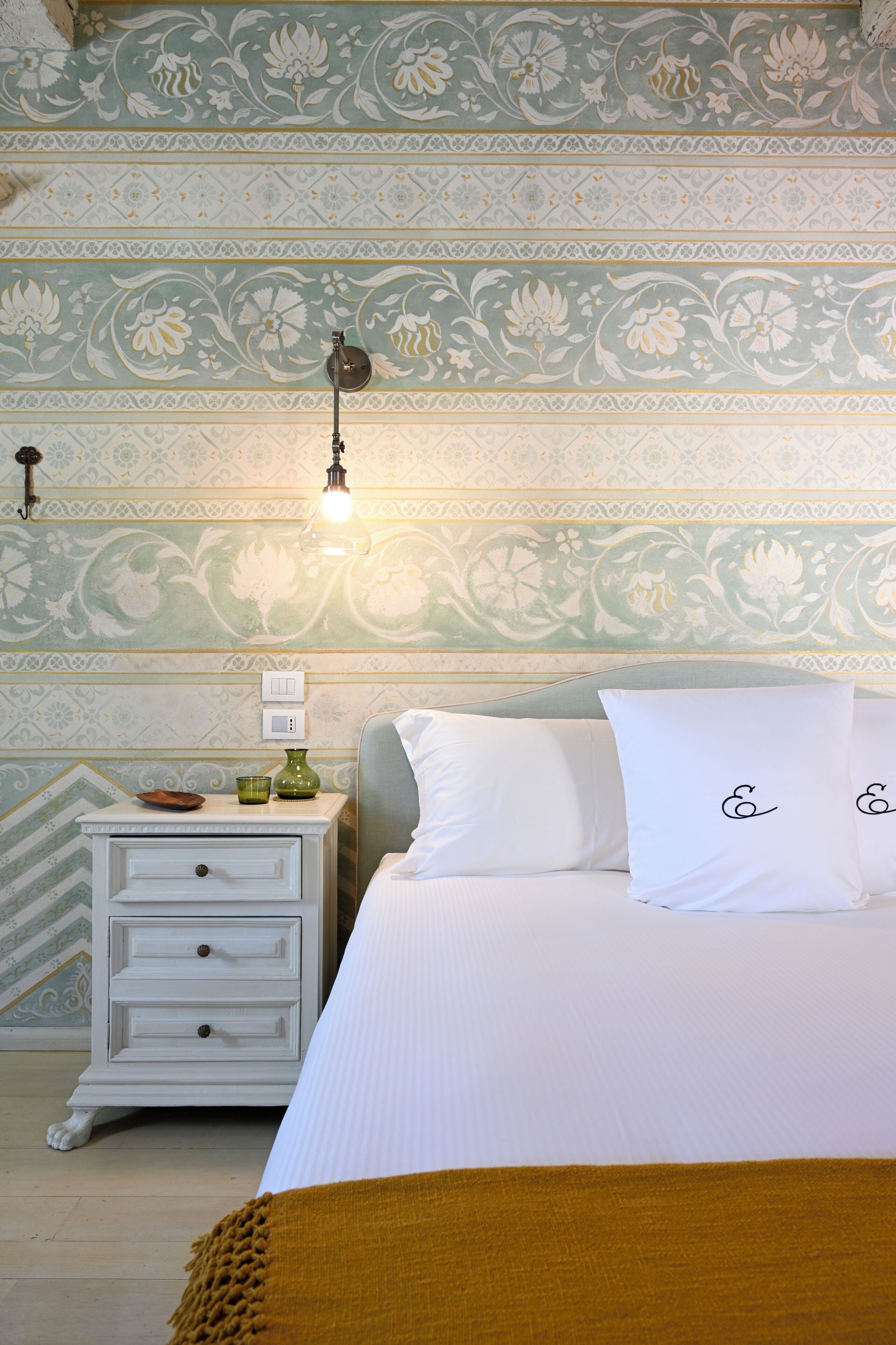 Une chambre avec un papier peint à motifs, une table de chevet avec une lampe, un lit avec une literie blanche et un plaid de couleur moutarde.