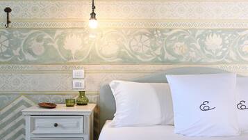 Une chambre avec un papier peint à motifs, une table de chevet avec une lampe, un lit avec une literie blanche et un plaid de couleur moutarde.