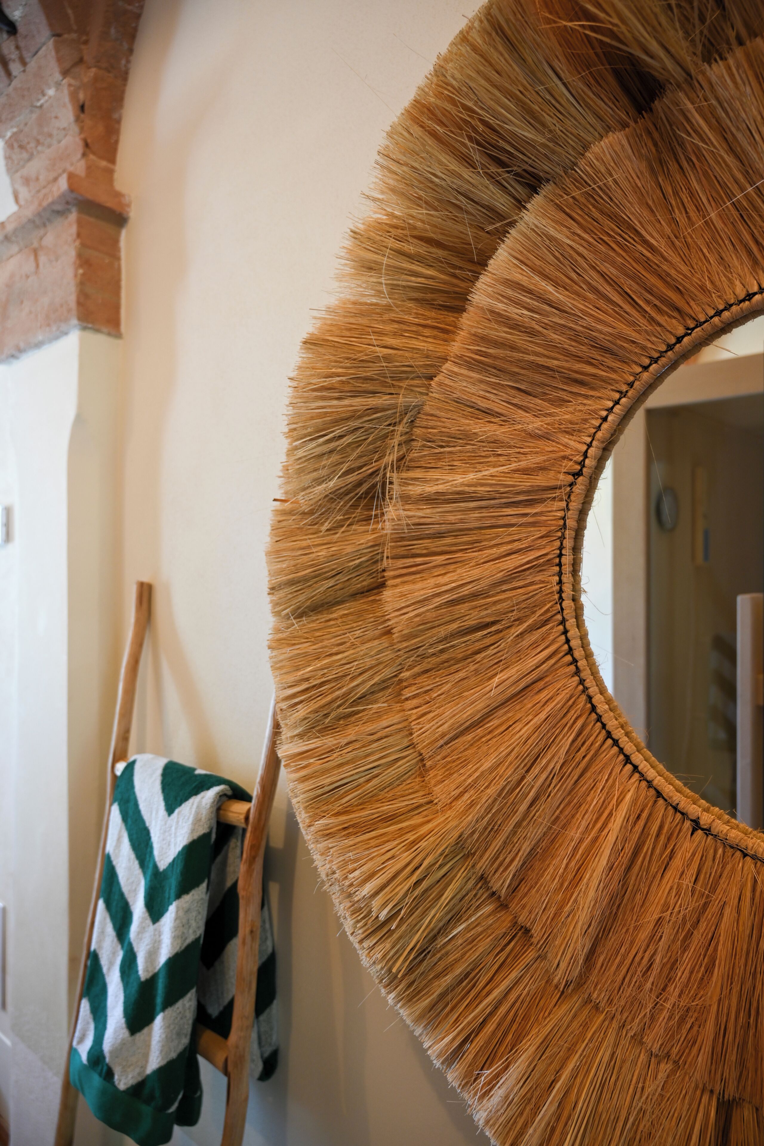 A round mirror with a straw-like texture, mounted on a wall with a brick arch. A wooden easel and a striped towel are visible in the background.